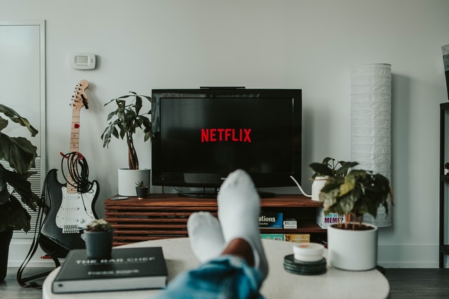 in einem Wohnzimmer sieht mand die Füße mit weißen Socken einer Person und dahinter ein Ferseher auf dem das Netflixlogo aufleuchtet