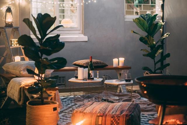 eine Terrasse mit einem weiß-schwarzen Teppich, zwei große Pflanzen, eine Holzbank mit Kissen, viele Kerzen und eine Lichterkette vor dem Fenster