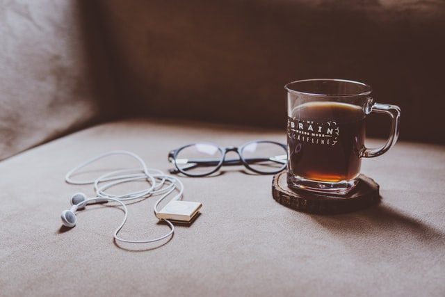 ein Glas mit Schwarztee auf einem Holzunterstzer, links daneben eine blaue Brille und links davon weiße Kopfhörer mit einem MP3-Player