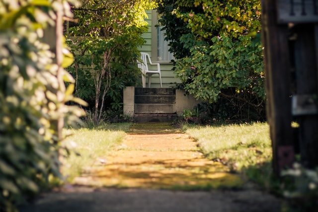 Der Garteneingang zu einem Haus, am Ende gibt es einige Treppen und oben auf der Terrasse steht ein weißer Plastikstuhl.