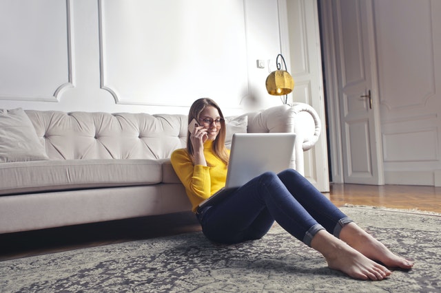 eine Frau in Jeans und gelben Pullover die auf einem grauen Teppich vor einem Sofa sitzt und telefoniert und einen Laptop auf den Beinen hat