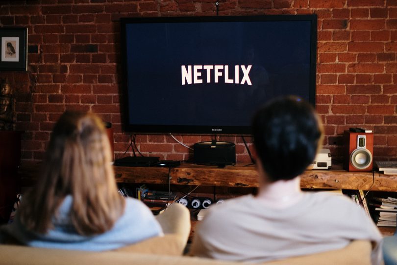 ein junges Pärchen das auf dem Sofa sitzt und Netflix in einem Fernseher an der Wand schaut