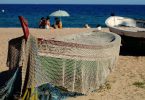 am Strande ein kleines Fischerboot, das mit einem Netz bedeckt ist, im Hintergrund siitzen drei Freunde im Sand unter einem Sonnenschirm und spielen Karten