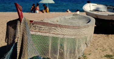 am Strande ein kleines Fischerboot, das mit einem Netz bedeckt ist, im Hintergrund siitzen drei Freunde im Sand unter einem Sonnenschirm und spielen Karten