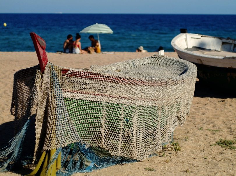 am Strande ein kleines Fischerboot, das mit einem Netz bedeckt ist, im Hintergrund siitzen drei Freunde im Sand unter einem Sonnenschirm und spielen Karten