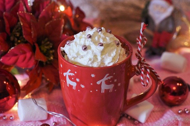 eine rote Tasse mit weißen Rentieren in dem heiße Schokolade mit Sahne oben drauf ist und die auf einem weihnachtlich geschmückten Tisch steht