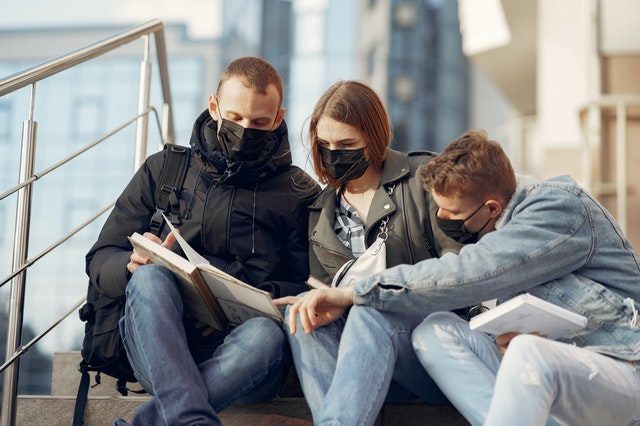 drei Studenten, zwei Männer und eine Frau in der Mitte, die alle schwarze Gesichtsmasken tragen, draußen auf einer Treppe sitzen und auf ein Buch schauen und zeigen