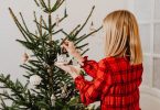 eine blonde Frau im kariertem rotem Hemd steht vor einem geschmücktem Weihnachtsbaum und hängt ein kleines weißes Häuschen dran