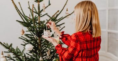 eine blonde Frau im kariertem rotem Hemd steht vor einem geschmücktem Weihnachtsbaum und hängt ein kleines weißes Häuschen dran