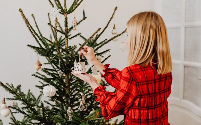 eine blonde Frau im kariertem rotem Hemd steht vor einem geschmücktem Weihnachtsbaum und hängt ein kleines weißes Häuschen dran