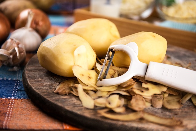 ein Holzbrett mit drei geschälten Kartoffeln, daneben liegt ein weißer Kartoffelschäler und dahinter mehrere Zwiebeln