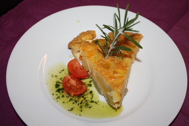 ein weißer runder Teller mit einem Stück spanischer Tortilla, in der Mitte steckt Rosmarien und daneben Olivenöl und zwei Stückchen Tomate