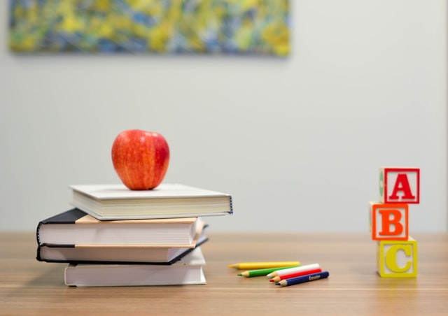 ein Holztisch auf dem vier Bücher aufeinander liegen und darauf ein roter Apfel, daneben mehrere Buntstifte und ganz rechts Würfel mit Buchstaben