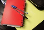 ein gelbes notizbuch auf dem ein rotes notizbuch auf dem ein blauer und ein schwarzer kugelschreiber liegen