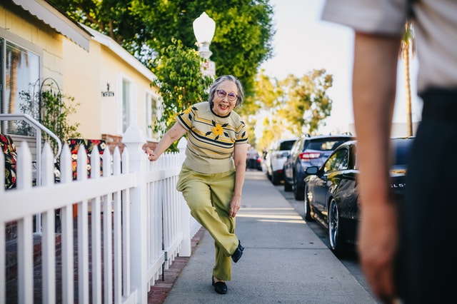 eine ältere Frau die sich an einem weißen Gartenzaun festhält um sich ihre grüne Hose zurechtzurücken